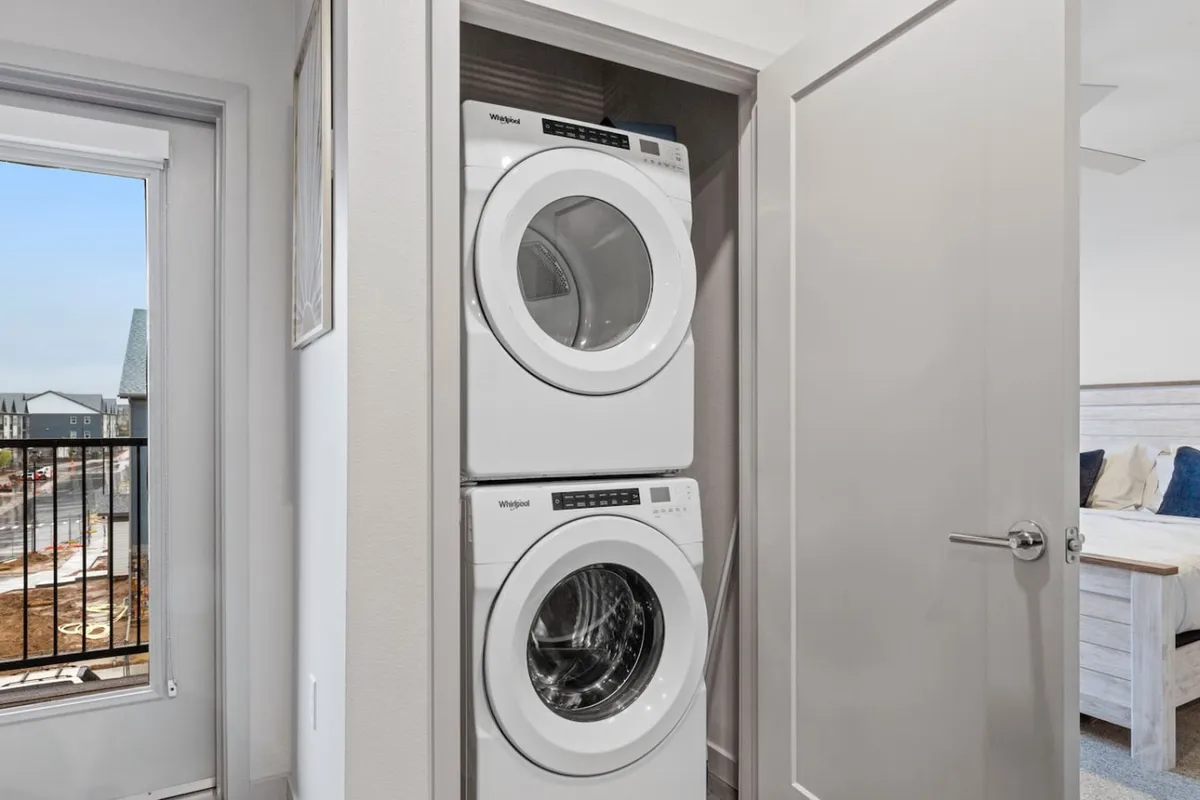 This image spotlights a modern laundry area with a stacked washer and dryer adjacent to a sunlit doorway leading to a balcony. Ample natural light enhances the practical and organized vibe, and the setup illustrates easy access to laundry amenities within the home.