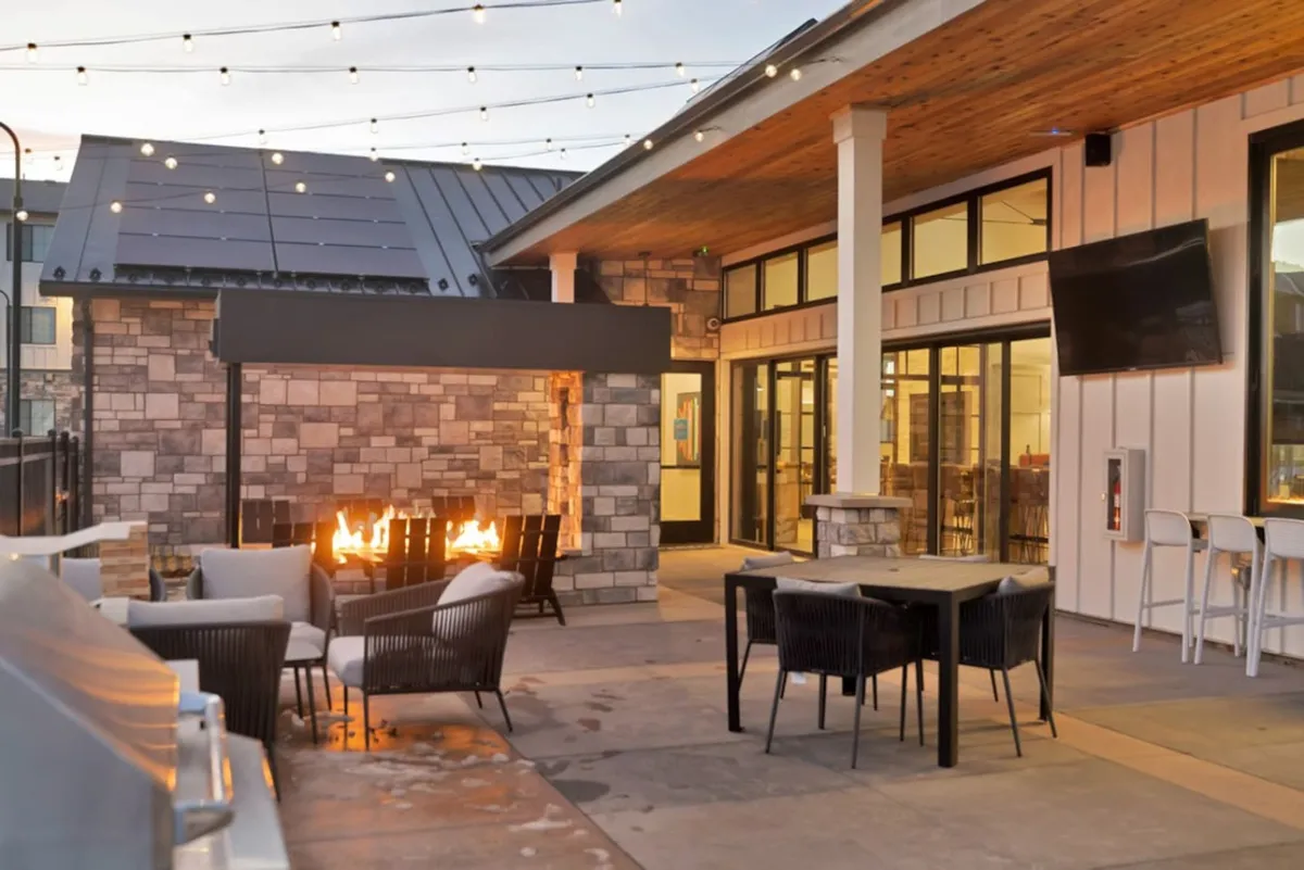 The image showcases the modern exterior of the apartment complex at dusk, with a welcoming fire pit surrounded by comfortable outdoor seating. Warm string lights create a cozy ambiance, inviting guests to gather, relax, and enjoy evenings under the Colorado sky.