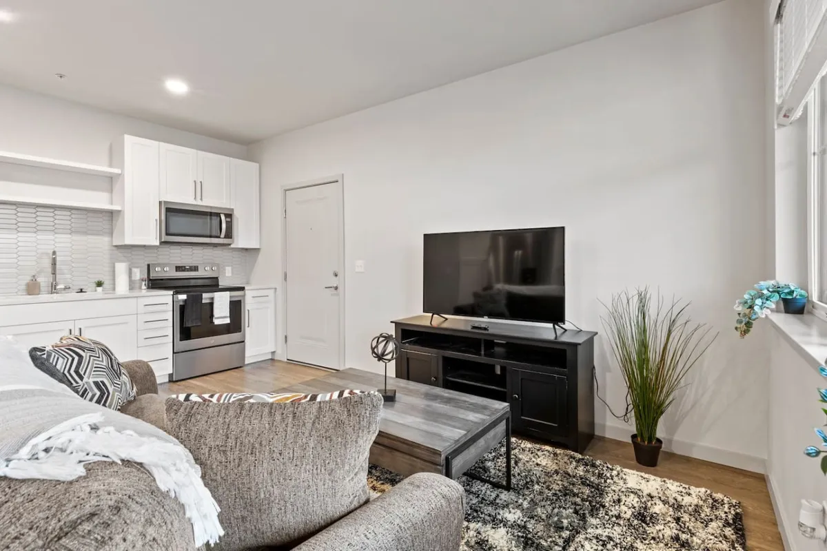 This interior photo captures a spacious open-plan living area featuring a comfortable sofa, television, kitchen area, and dining table. The hardwood-style floors and plush furnishings are visible, with natural light streaming through the windows, creating a cozy and inviting atmosphere.
