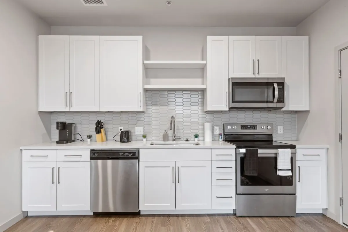 A stylish, contemporary kitchen featuring pristine white cabinetry, sleek stainless steel appliances—including a prominent refrigerator and oven—a striking hexagonal tile backsplash, and a deep kitchen sink. The space is bright and airy, inviting both daily meal prep and special culinary adventures.
