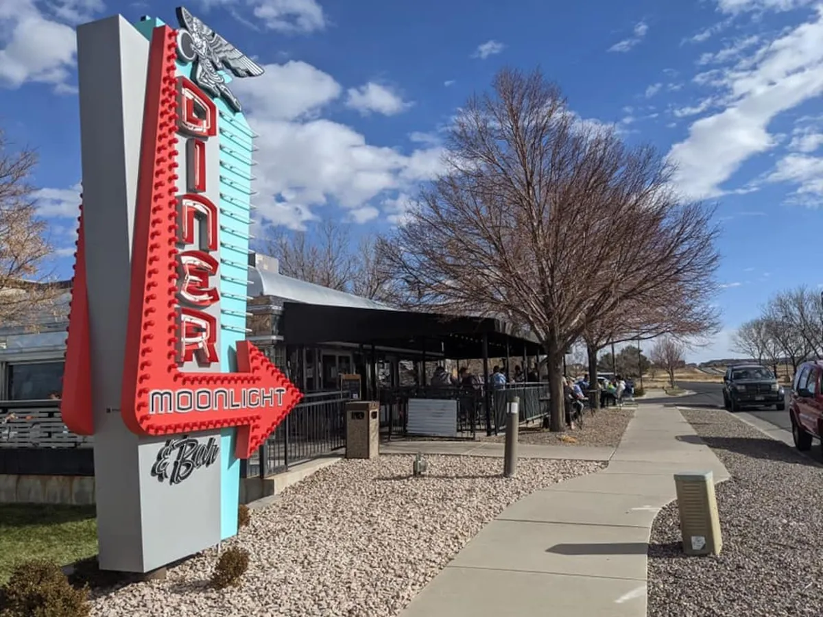 An exterior scene featuring a neon diner sign, outdoor seating area, and a sidewalk bustling with activity. The image captures a lively street atmosphere, perfect for socializing, dining, and enjoying the neighborhood vibe.