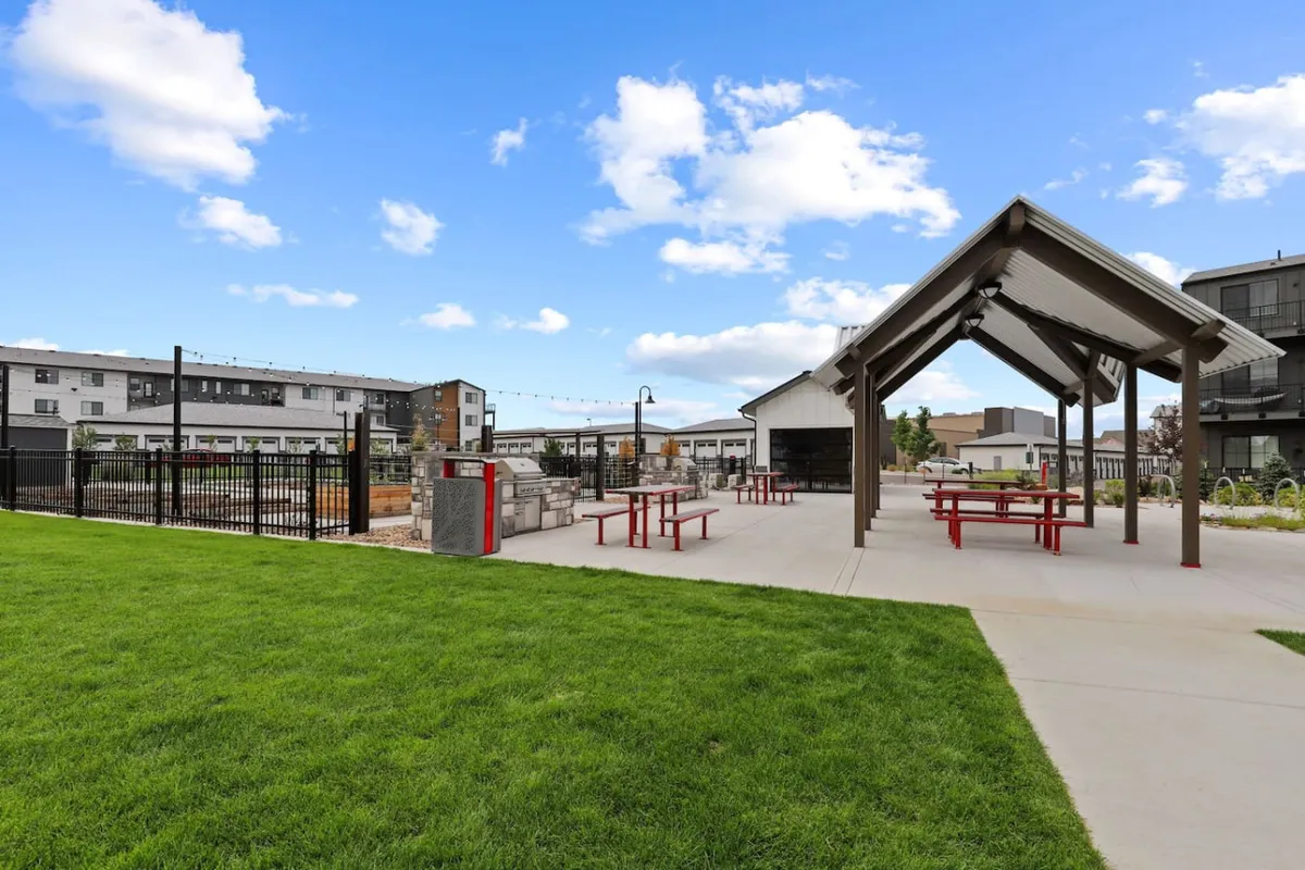 This exterior image captures a cheerful community pavilion with a roof, red picnic tables, a green lawn, and a fenced garden area. Residents are seen enjoying picnicking, community gatherings, and outdoor games, creating a lively, social atmosphere. The setting is perfect for memorable barbecues, holiday celebrations, and neighborhood events.